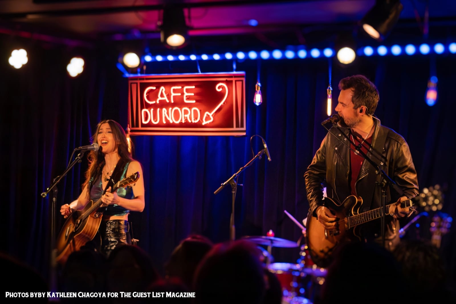 Two guitarists singing under the neon sign, in front of a blue curtain