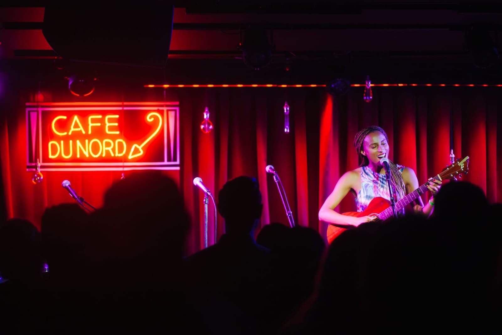 A singer with a guitar on the Cafe du Nord stage under the red neon sign, seen over the heads of the crowd
